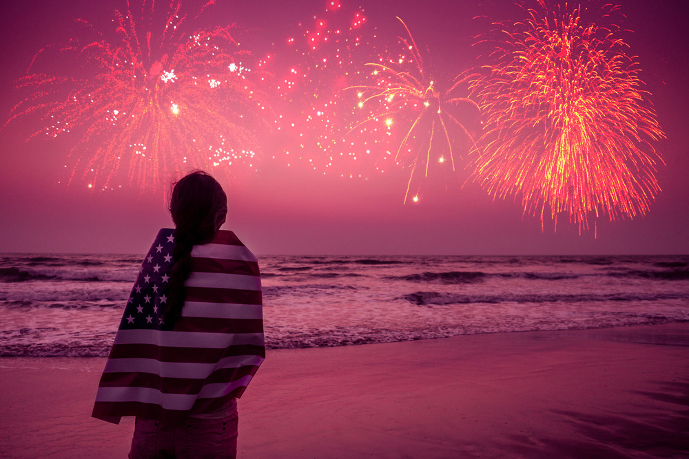 A woman wrapped in an American flag watches fireworks over the water at the beach. Attendance of one of the best 4th of July events in Florida.